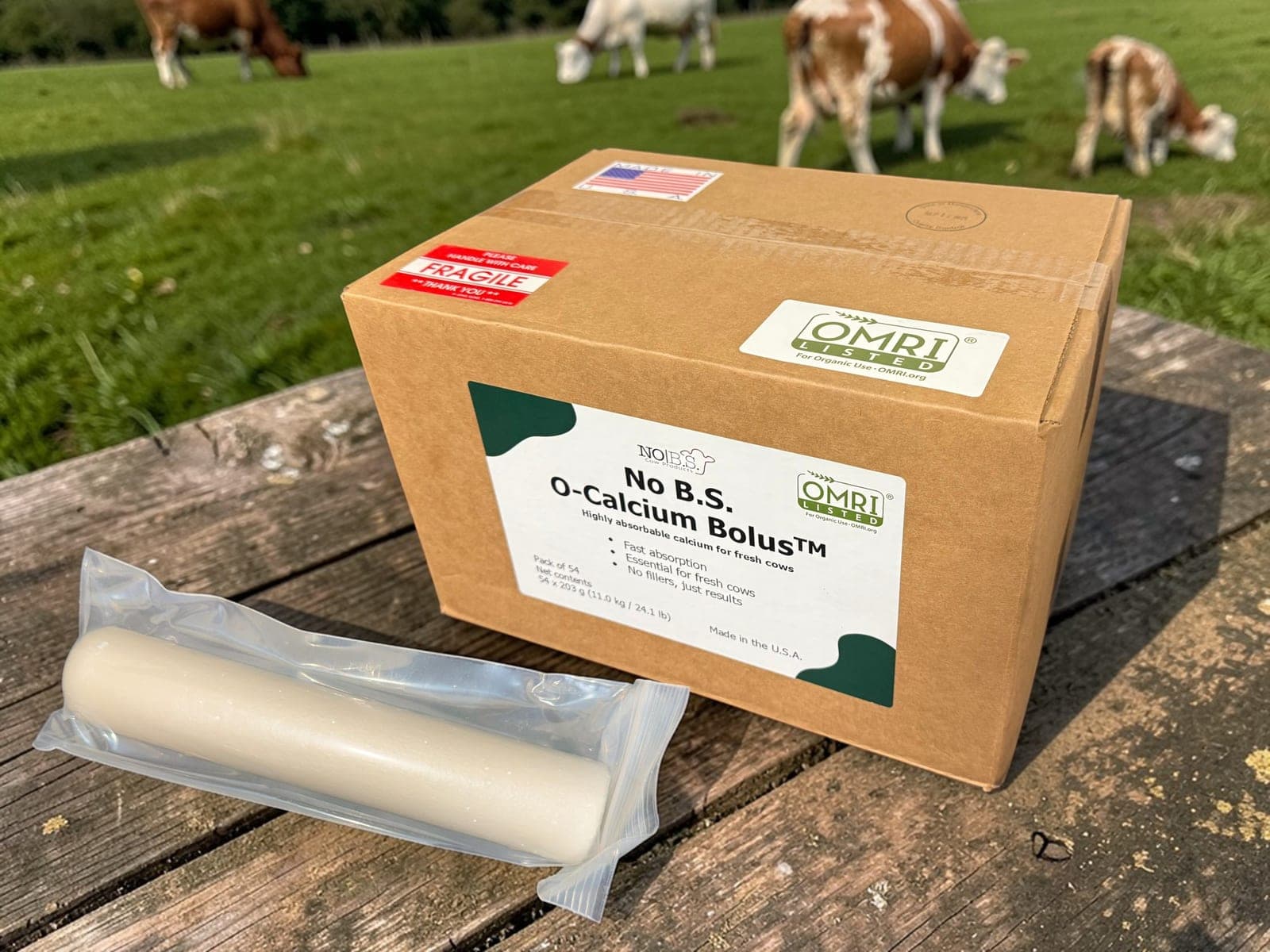 A case of OMRI Listed No B.S. O-Calcium Bolus and a single sealed bolus on a bench in a pasture.